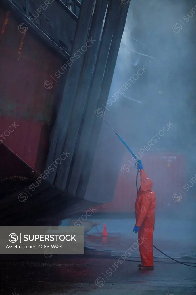 A crew member washes boat hull using high_pressure hose and nozzle while vessel rests on keel blocks and in Marine Travelift slings, Kodiak Boat yard,...
