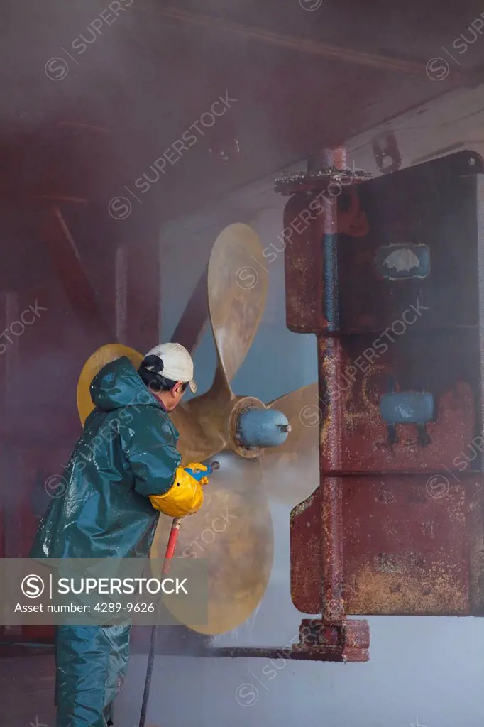 Crew member washing the propeller and stern of commercial fishing vessel using high_pressure hose and nozzle while boat rests on keel blocks and in Ma...