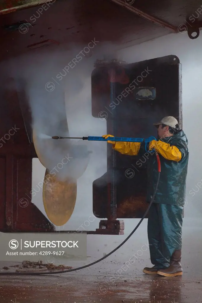 Crew member washing the propeller and stern of commercial fishing vessel using high_pressure hose and nozzle while boat rests on keel blocks and in Ma...