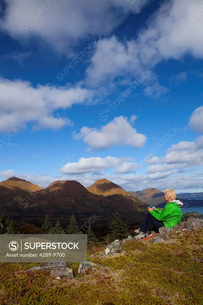 Mature woman relaxes and enjoys a hot drink on Pillar Mountain with Monashka Bay in background, Kodiak Island, Southwest, Alaska, Autumn