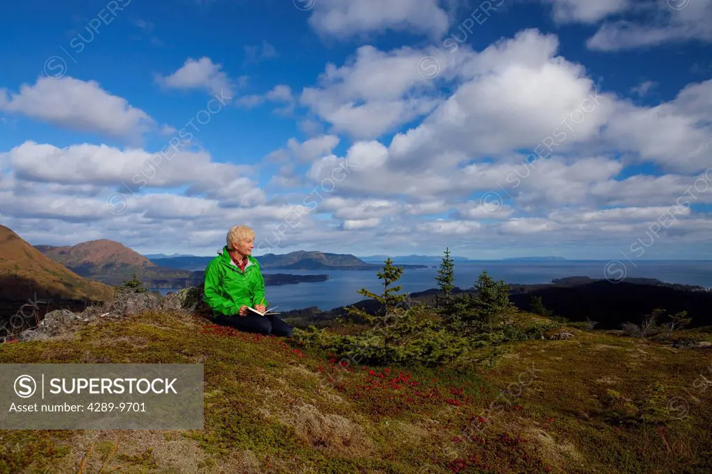 Mature woman relaxes and writes in journal on Pillar Mountain with Monashka Bay in background, Kodiak Island, Southwest Alaska, Autumn