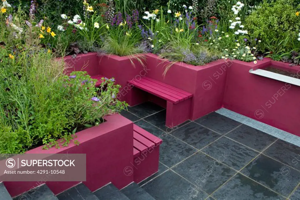 Bright pink walls and pink benches in sunken patio with slate paving and summer flowering plants in borders