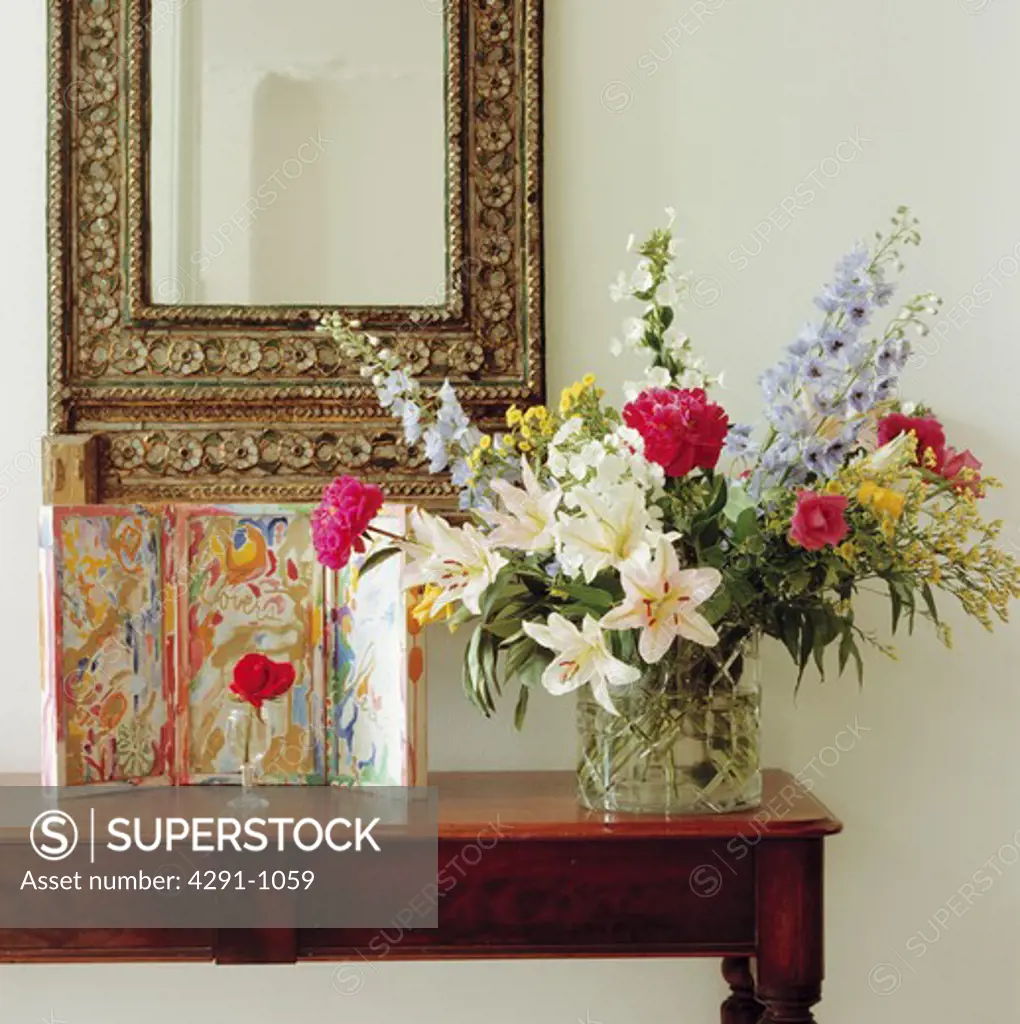 Close-up of small picture and summer flowers in informal arrangement in glass vase on table below ornate mirror