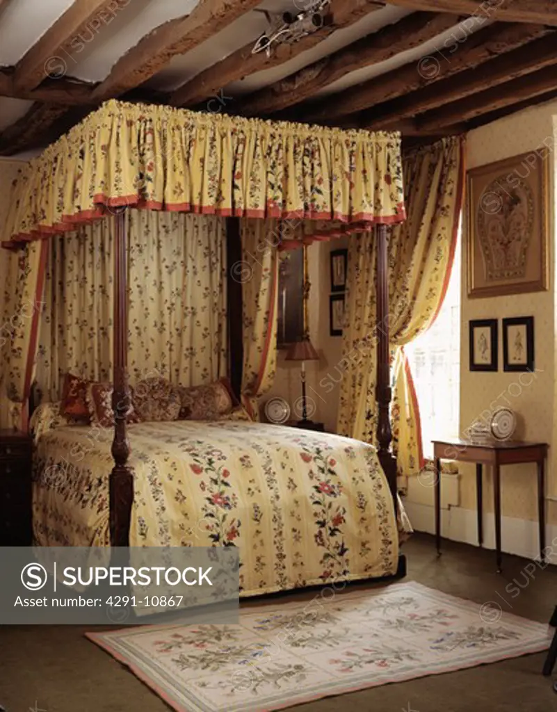 Antique four-poster bed with yellow floral quilt and drapes in traditional country bedroom with floral rug and beamed ceiling