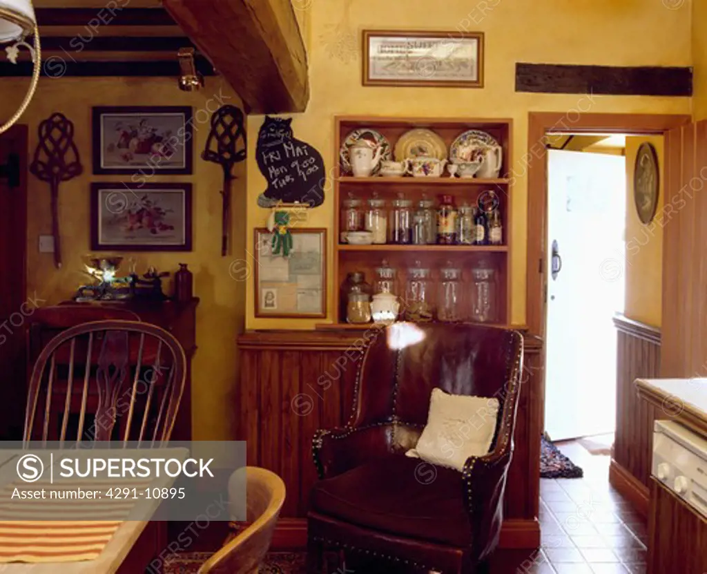Brown leather armchair in front of alcove shelves with glass jars and pottery in yellow cottage kitchen
