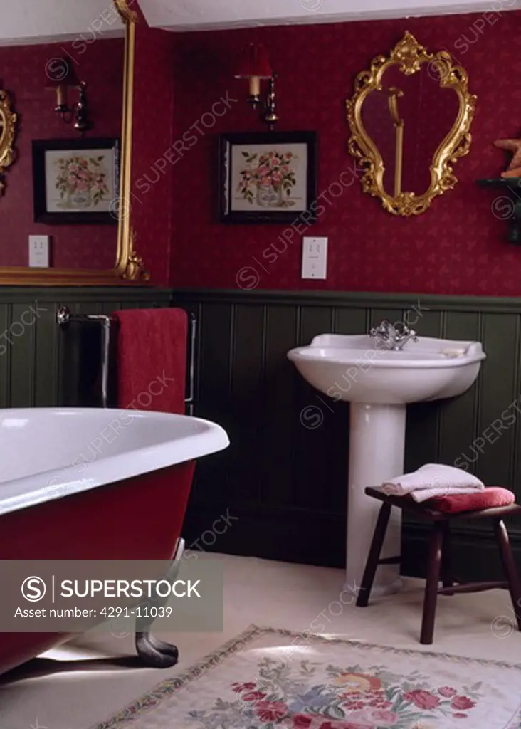 Ornate gilt mirror above white pedestal basin in red bathroom with green dado panelling and central rolltop bath