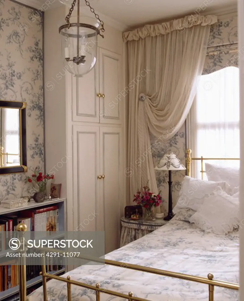 White curtains and blue and white blind above brass bed in bedroom with fitted white wardrobe and blue floral wallpaper