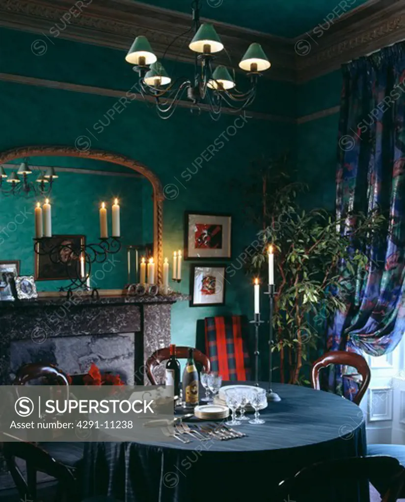 Gilt mirror above lighted candles on mantelpiece in green dining room with red tartan throw on chair