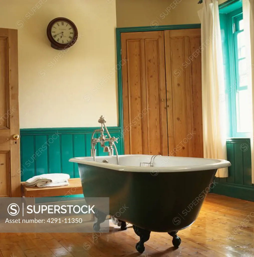 Roll-top bath on wooden flooring in bathroom with large fitted storage cupboard and painted green dado panelling