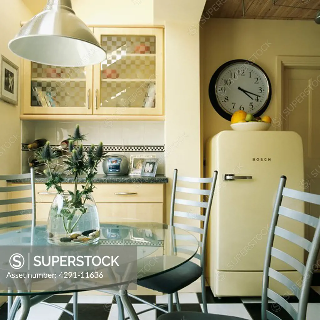 Circular glass table and metal chairs in cream modern kitchen with circular clock above cream freestanding Bosch fridge