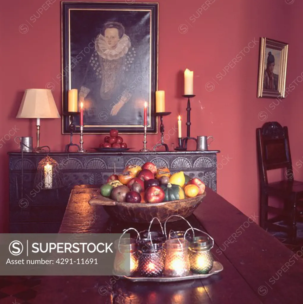 Candles in glass jars on table with gourds on plate in red dining room with oil painting and candles on sideboard