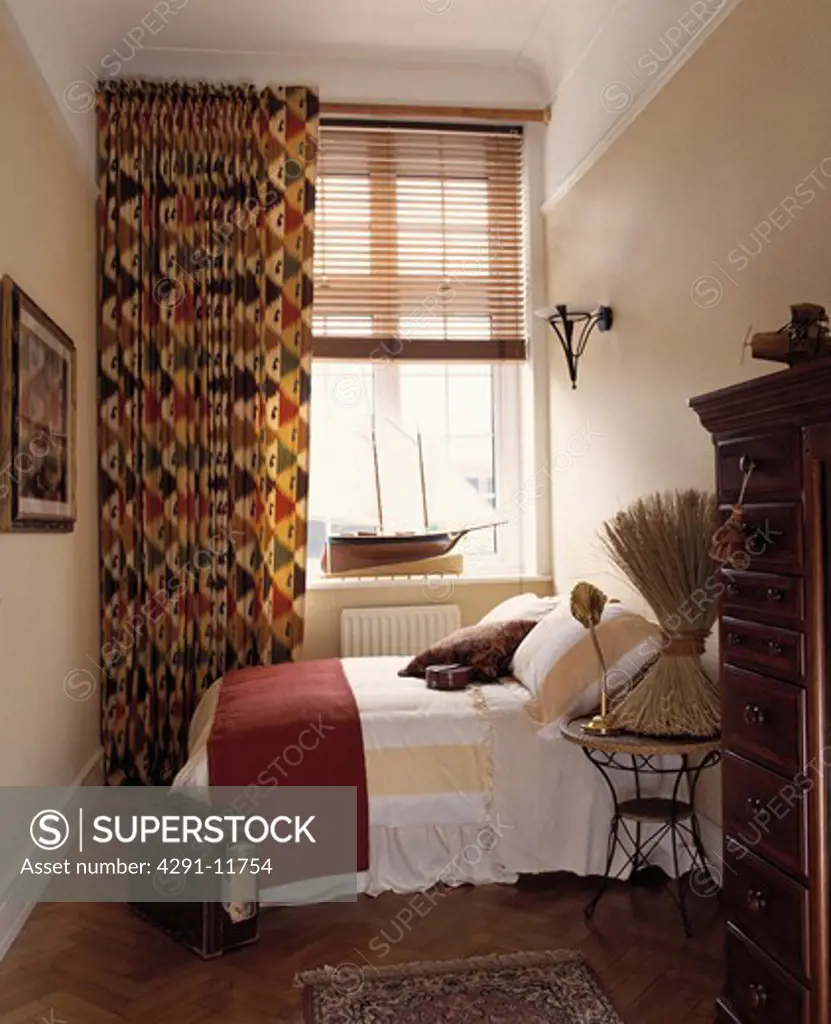 Patterned red and green curtains and slatted wooden blind at window in narrow white bedroom