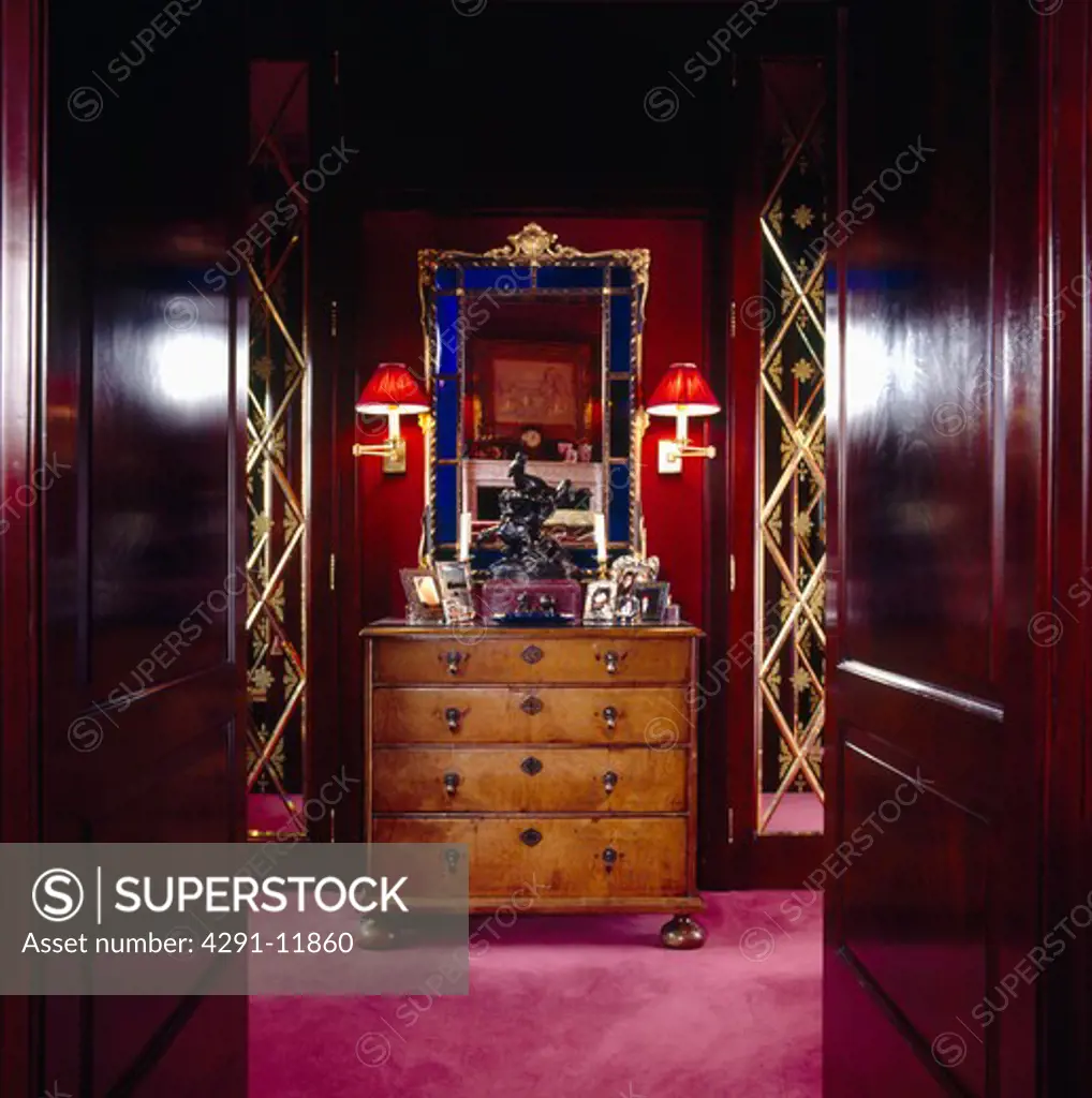 Double mahogany doors open to dressing room with ornate blue and gold framed mirror above antique chest-of-drawers