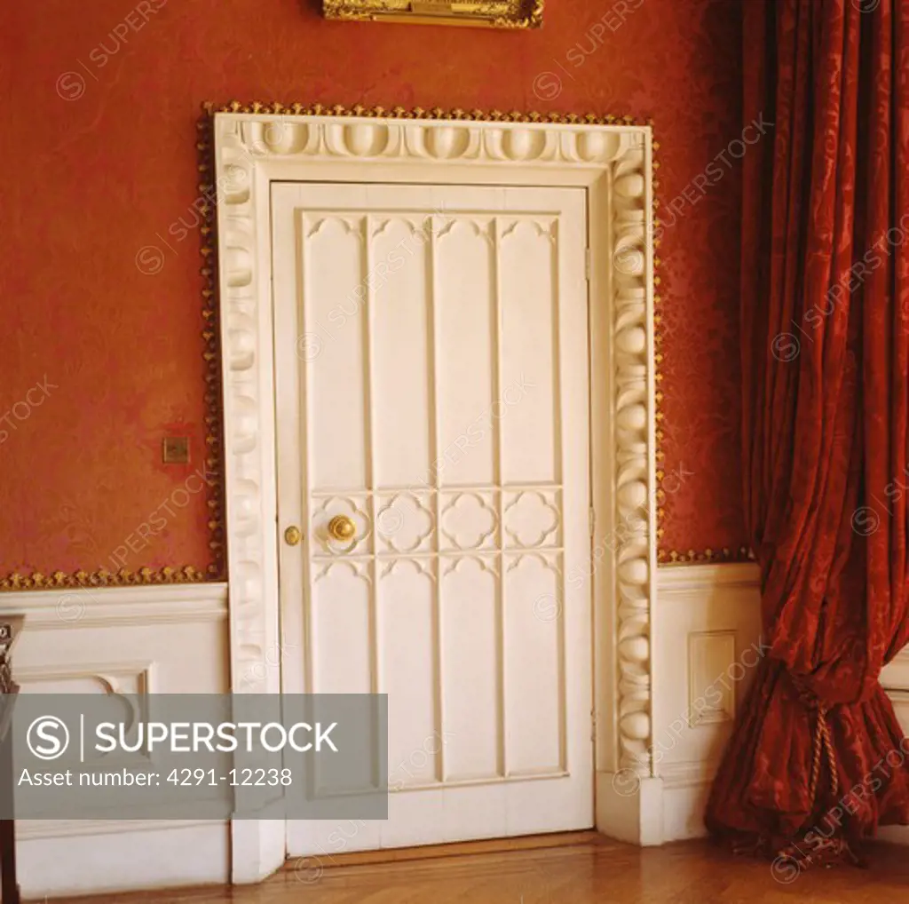 Close up of white Gothic door with ornately carved frame in red room with white wainscot panelling and red curtain