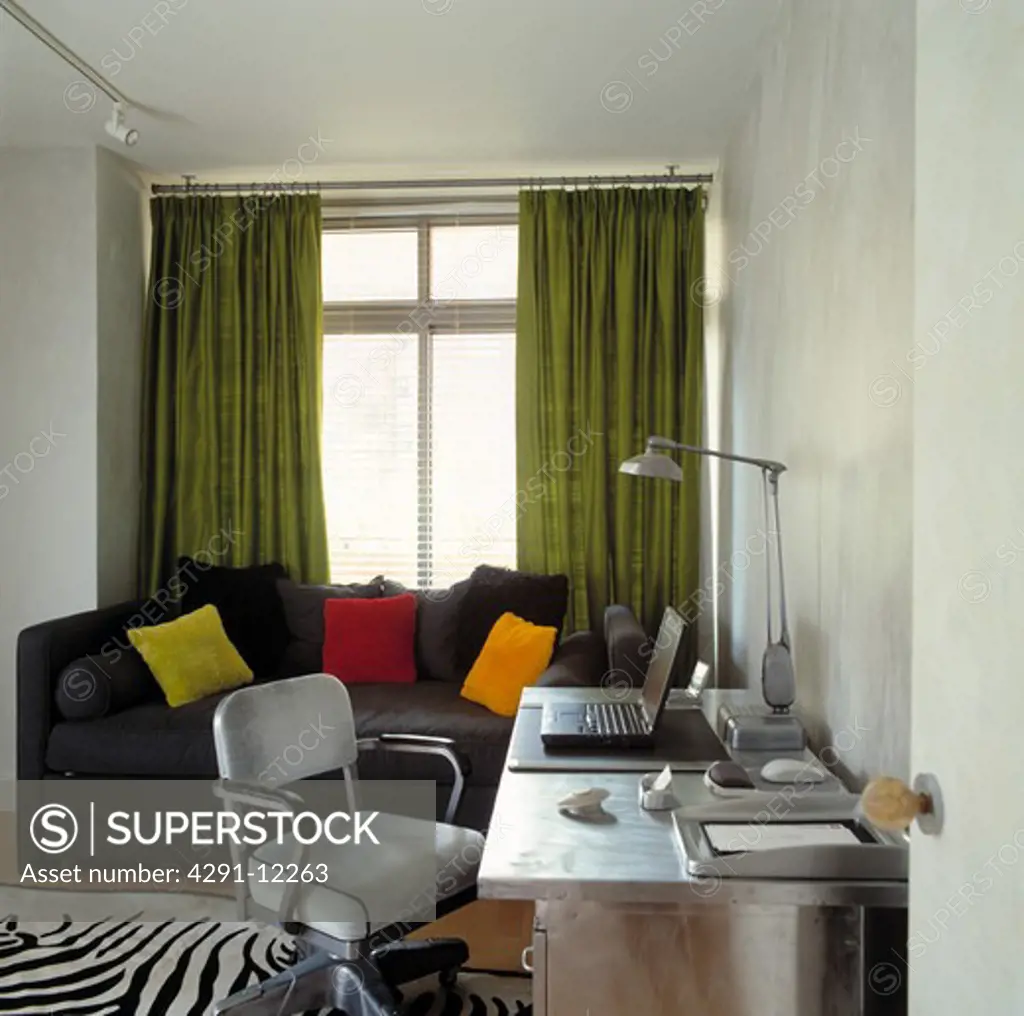 Laptop on stainless steel desk in small home office with sofa in front of window with green curtains