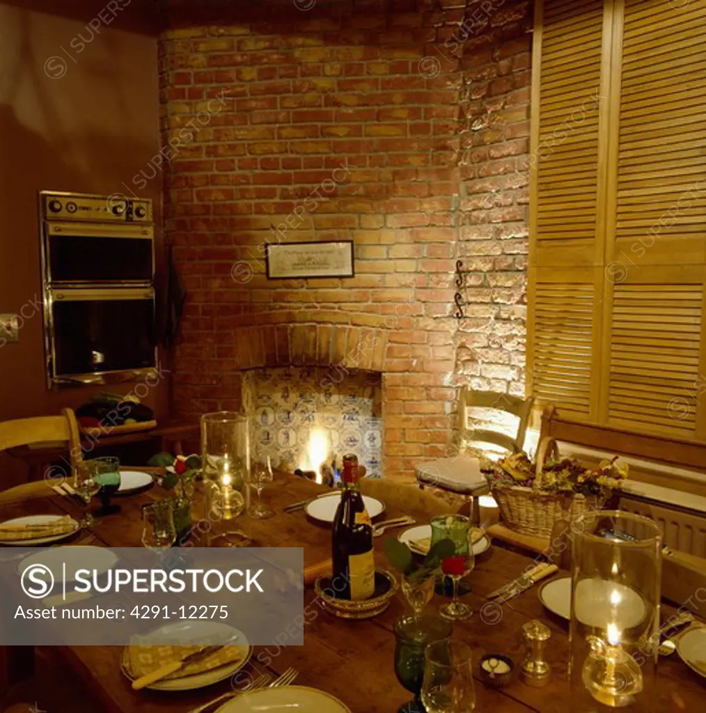 Place settings and lighted candles on wooden table in kitchen with fireplace in brick wall