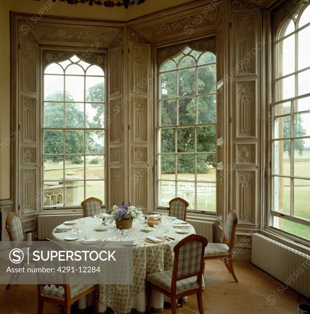 Patterned cloth on circular table in large dining room with tall Gothic windows and view of outdoors