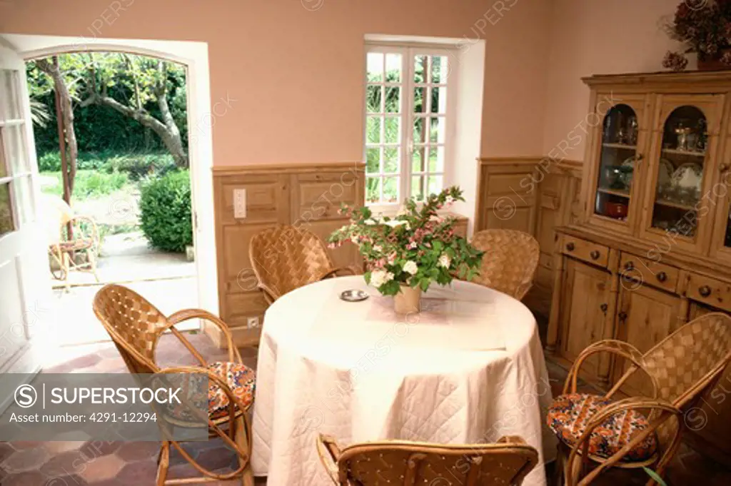 White quilted cloth on circular table with woven rattan chairs in country dining room with open door to garden