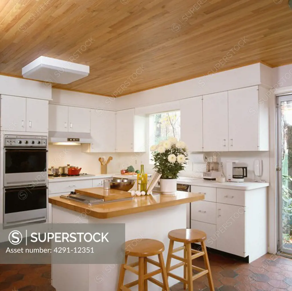 Wooden stools at island unit with wooden worktop in modern white kitchen with wooden ceiling