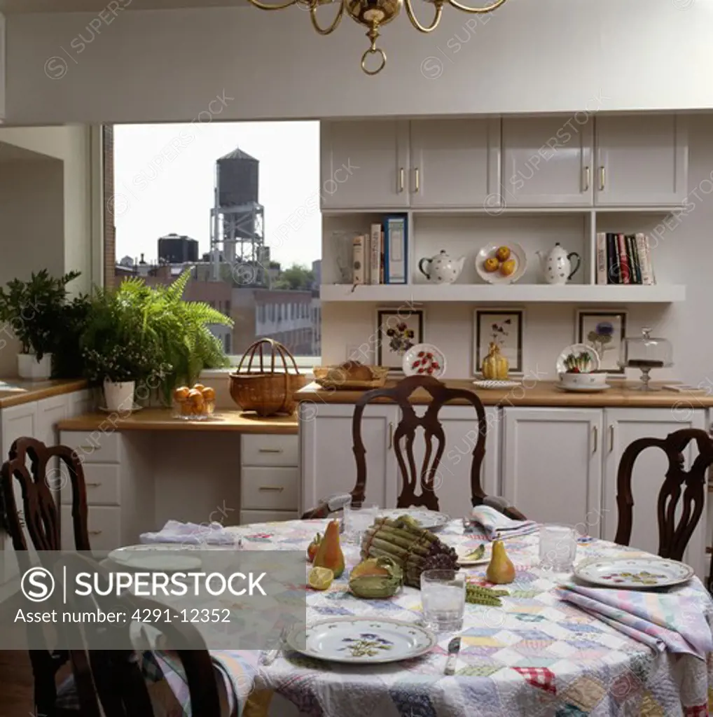 Place settings on patchwork cloth on circular table in white kitchen with view of city through window