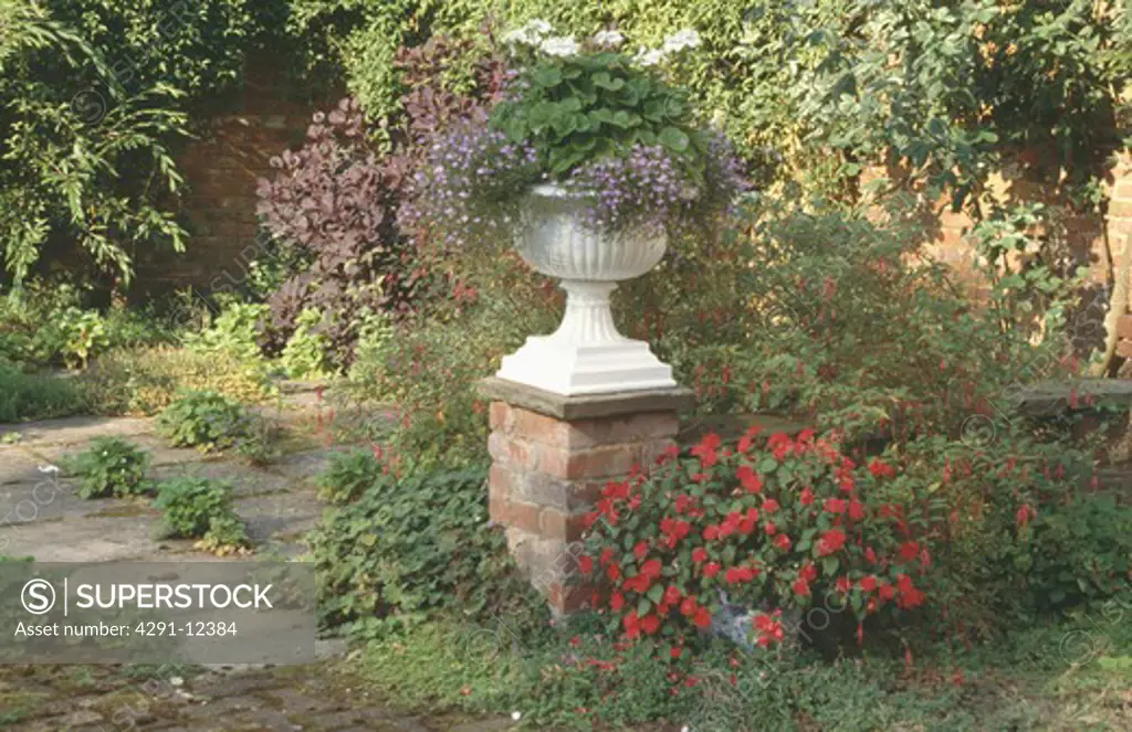 White urn on wall above red impatiens in pot in country garden