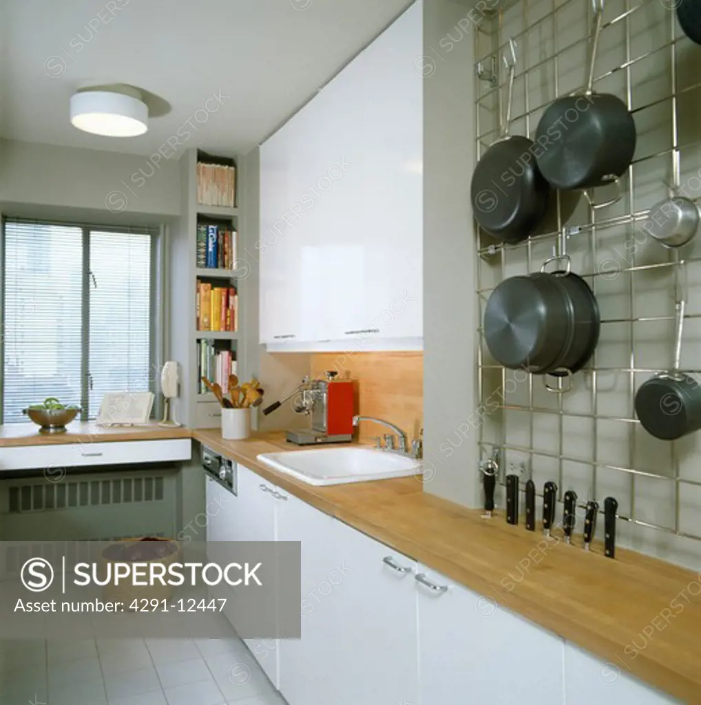 Pans on metal wall-mounted storage rack in modern white galley kitchen with wood worktop and white sink