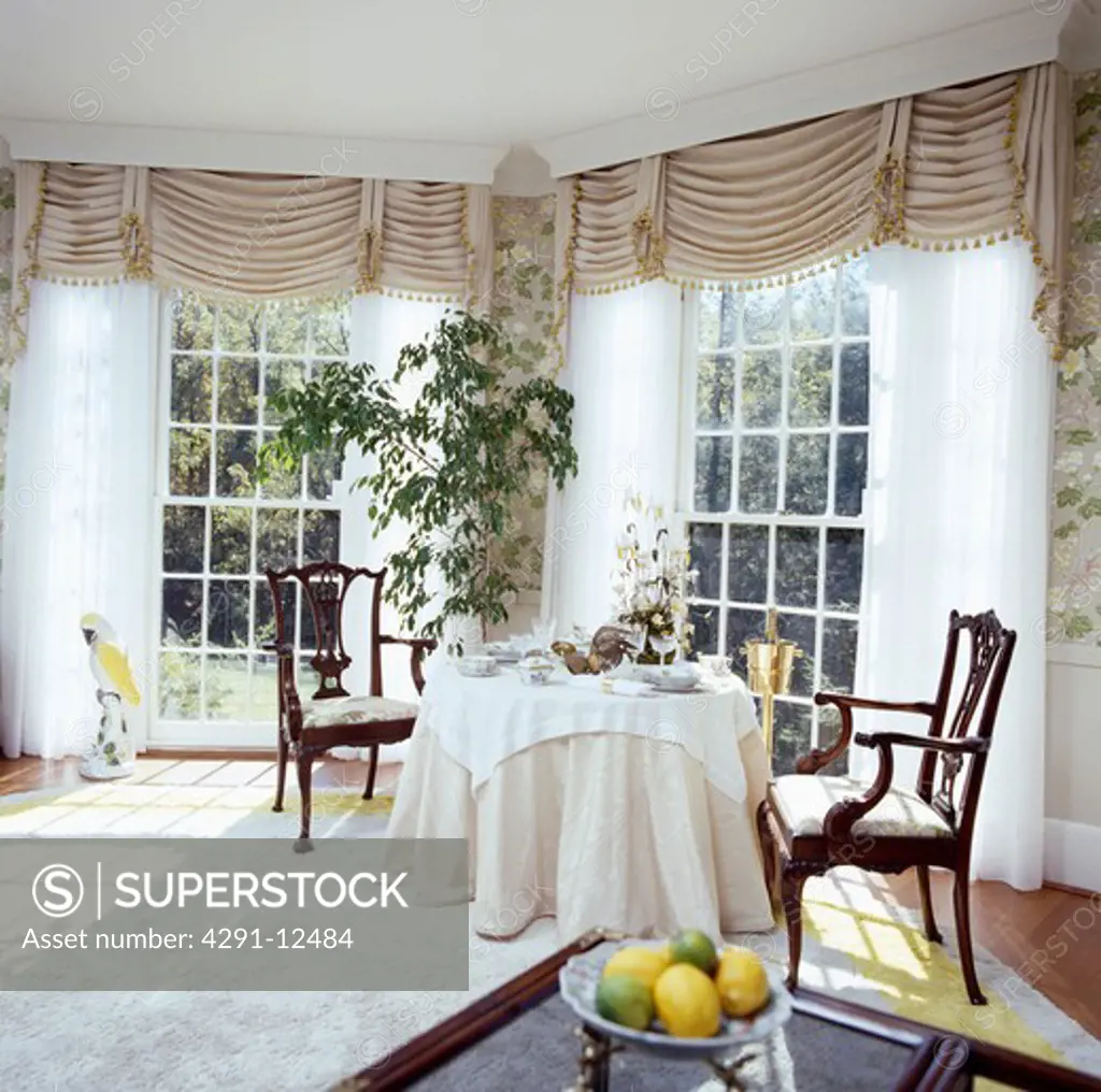 Beige ruched blinds and white curtains at tall windows in eighties diningroom with beige cloth on circular table