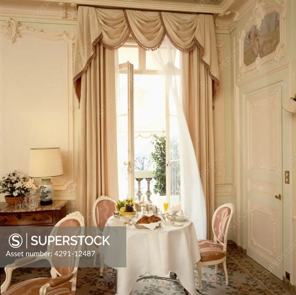 Table with white cloth set for breakfast in front of French windows with beige swagged curtains in chateau dining room
