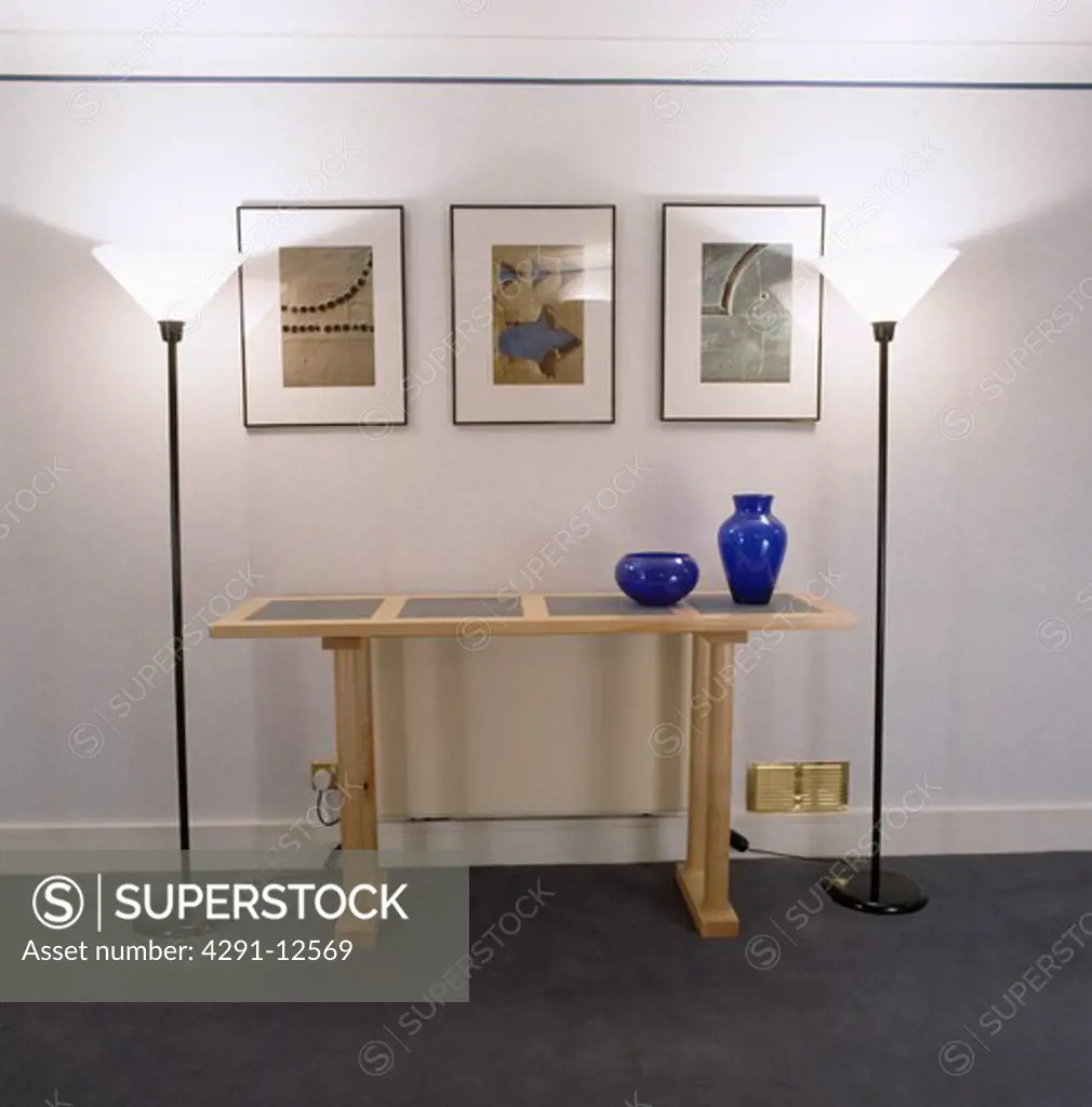 Pictures above pale wood modern console table in modern white hall with uplighter lamps