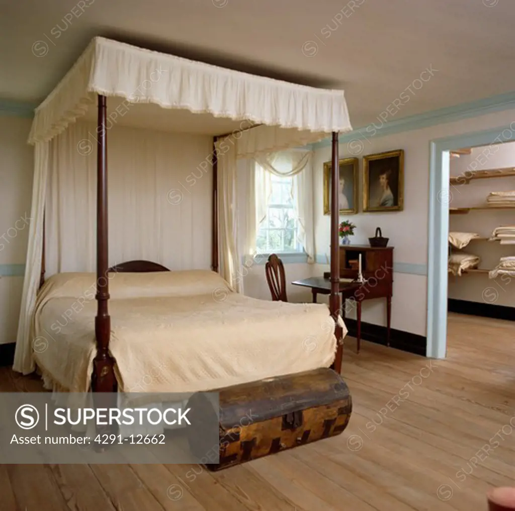 Small antique wooden chest in front of four-poster bed with cream bedcover in colonial-style bedroom with wooden floor