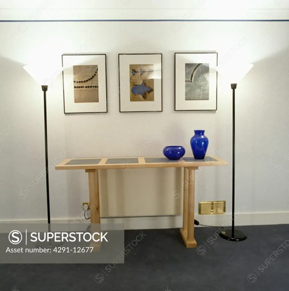 Pictures above pale wood modern console table in modern white hall with uplighter lamps