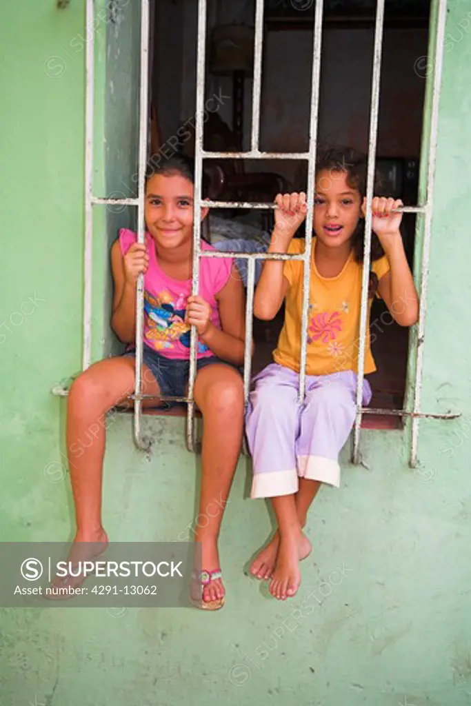Two young girls posing on a window sill, behind protective iron bars, Camaguey, Camaguey Province, Cuba