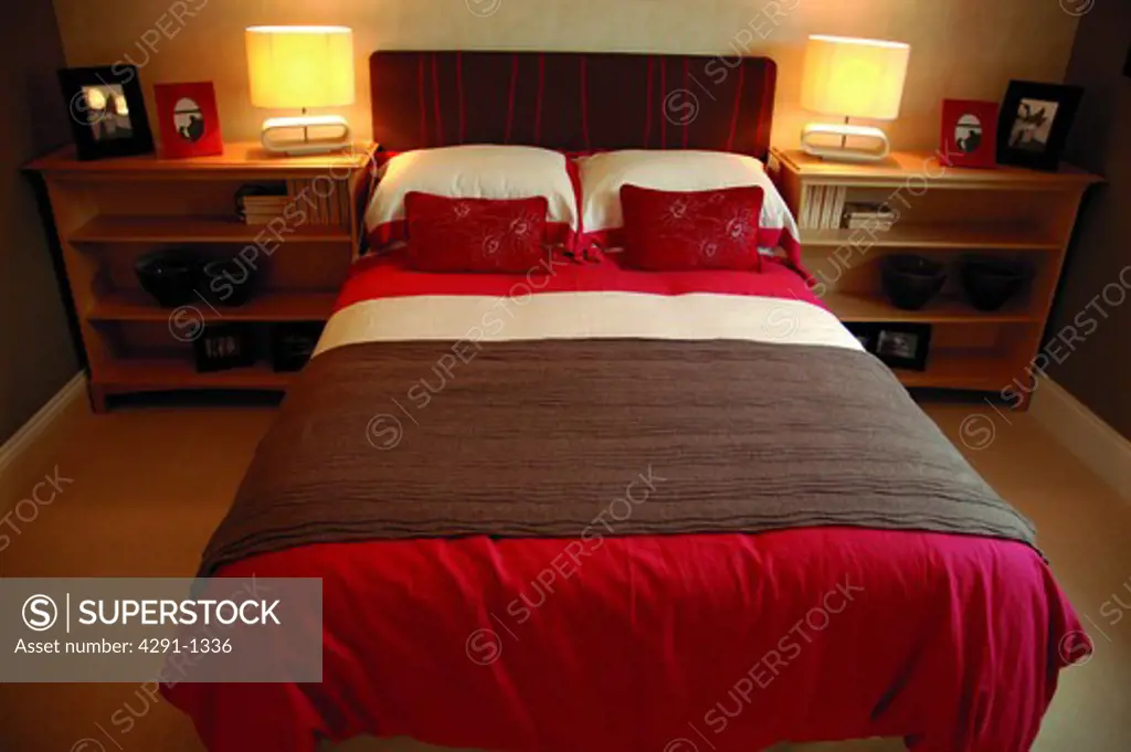 Lighted lamps on bedside tables on either side of bed with brown throw on red and white quilt in modern bedroom