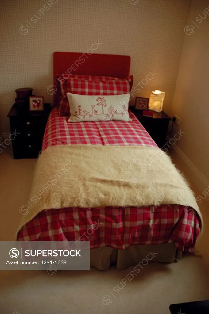 Cream mohair throw and patterned red duvet and pillows on single bed in modern bedroom with lighted lamp on table