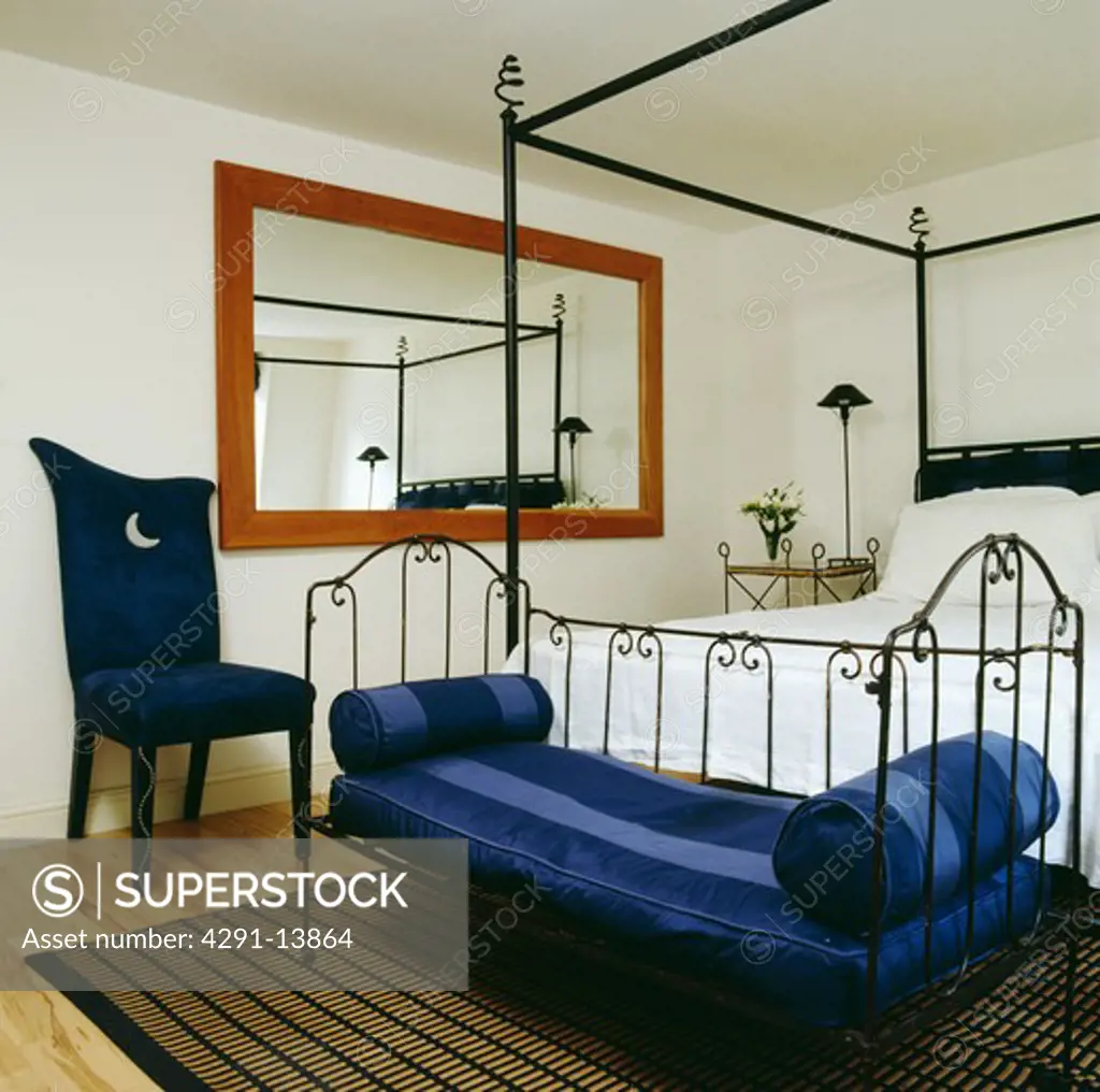 Blue striped cushions on metal daybed below modern black iron four-poster bed in white bedroom with blue chair