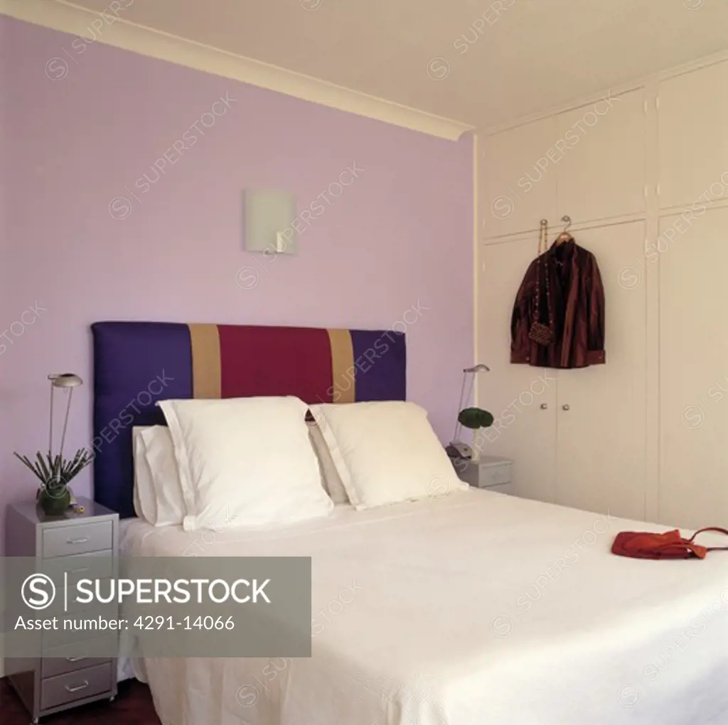 Mauve wall behind red blue and gold striped upholstered bed with white pillows and linen in bedroom with fitted wardrobe