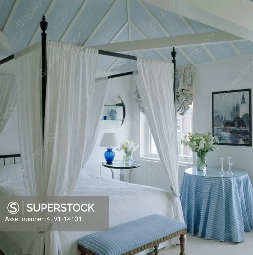 Bed with white drapes and linen in white bedroom with blue stool and cloth on table