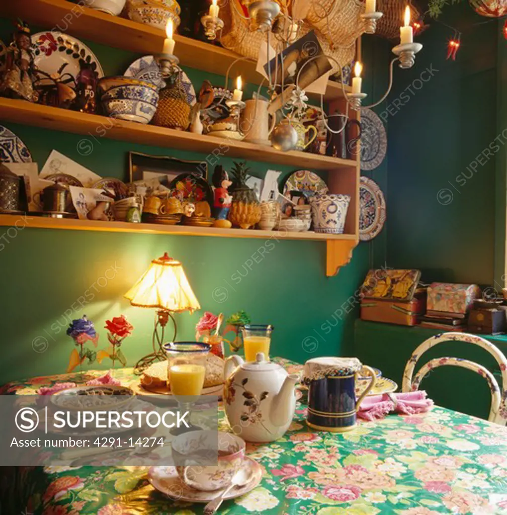 Green dining room with lighted lamp and old teaset on table with green floral cloth below shelves with collection of pottery