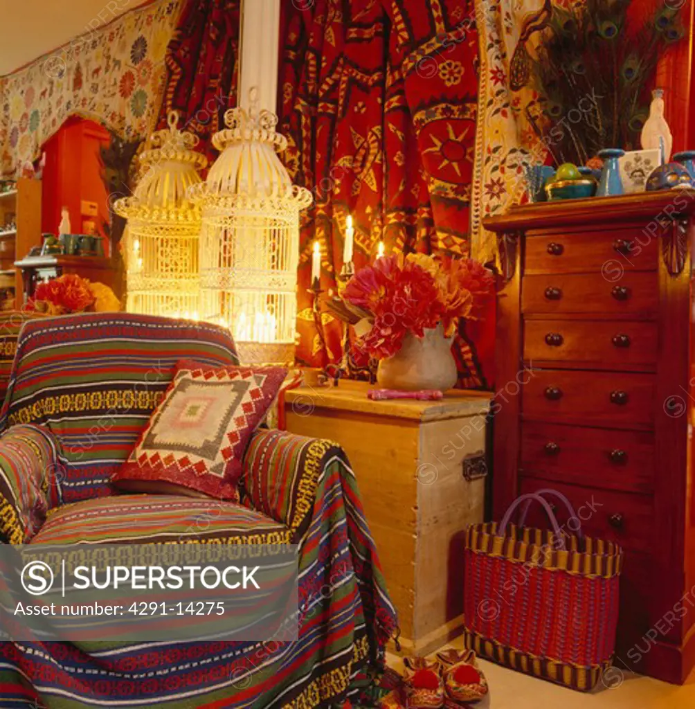 Striped ethnic throw and cushion on armchair beside wooden chest with lighted candles in ornate white birdcage