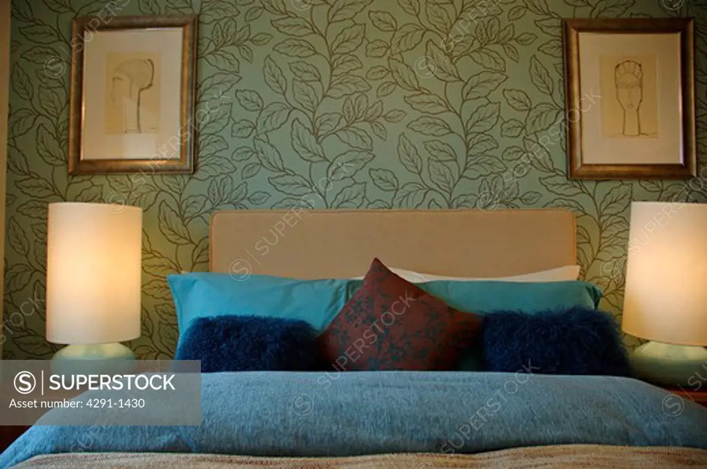 Lighted cylindrical lamps on either side of bed with blue pillows and cover in modern bedroom with green leaf-pattern wallpaper