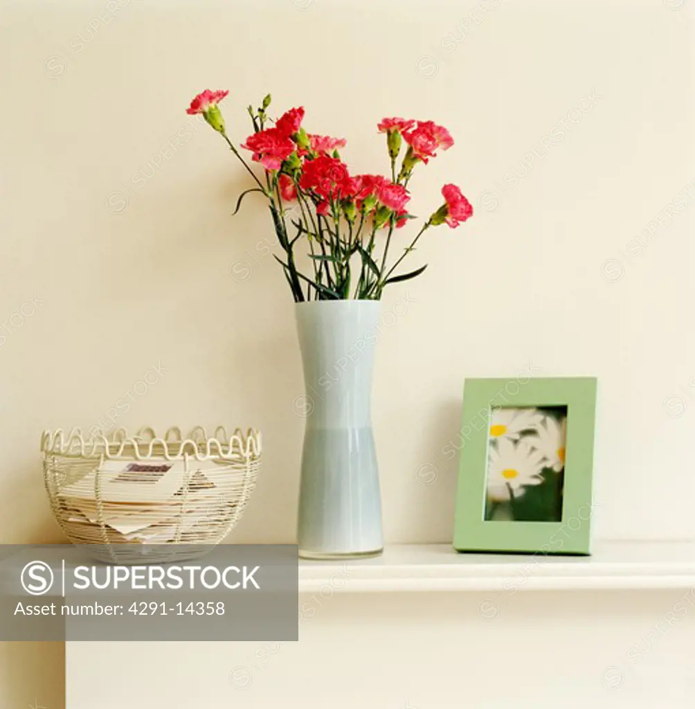 Close-up of vase of red flowers on mantelpiece with small picture and cream wire basket