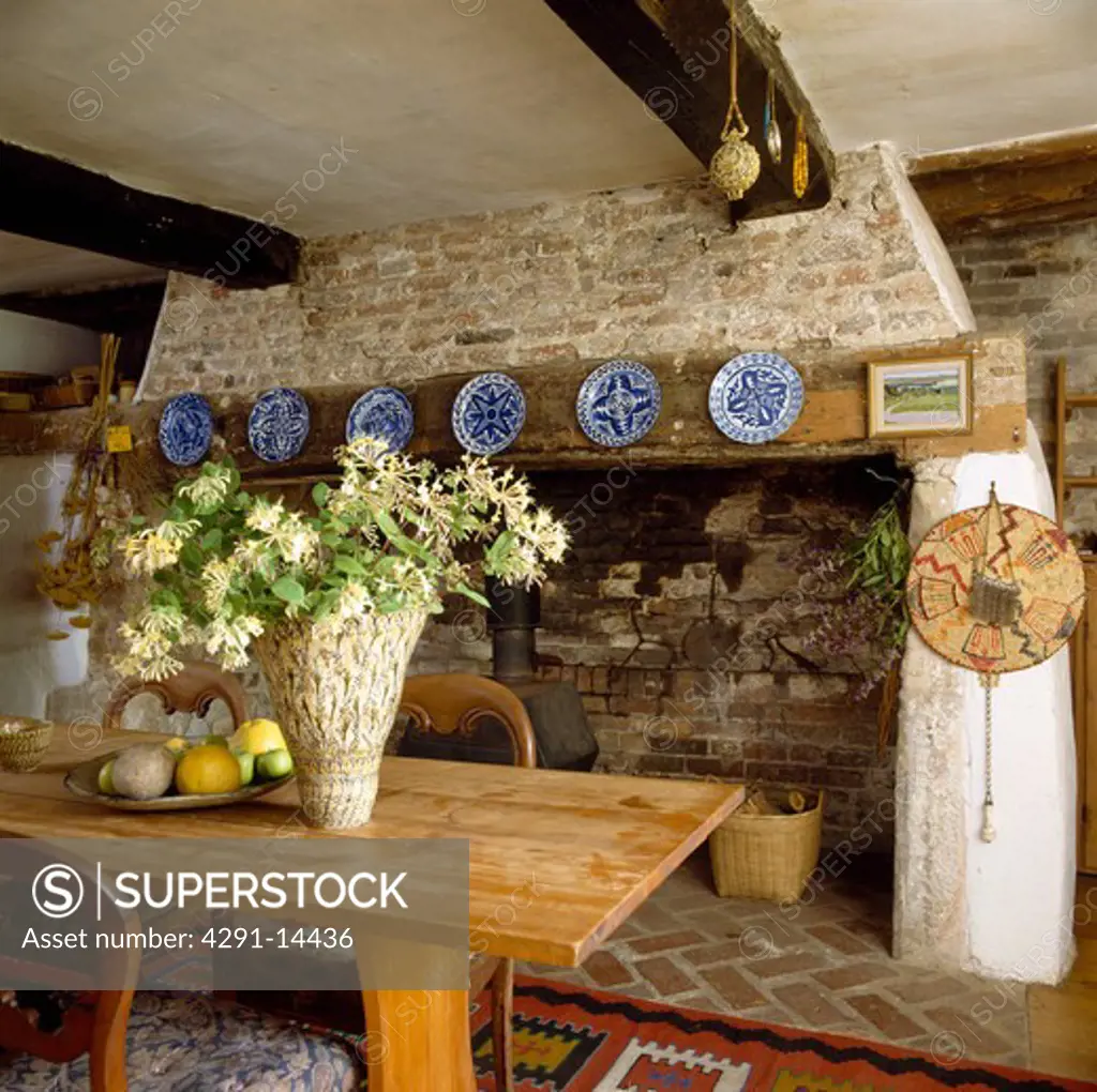 Pottery vase of honeysuckle on simple wood table in cottage dining room with inglenook fireplace