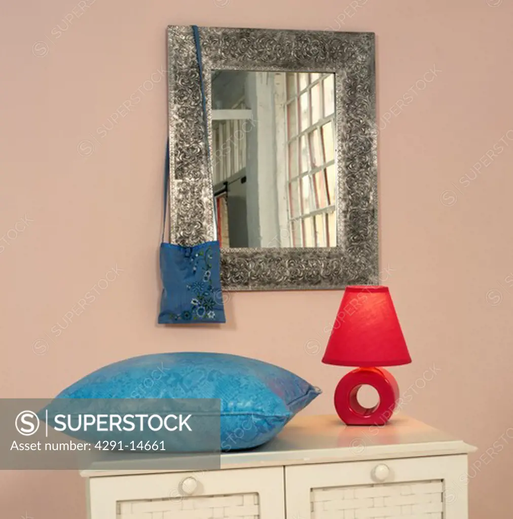 Close-up of metal-framed mirror above blue cushion and red lamp on chest-of-drawers