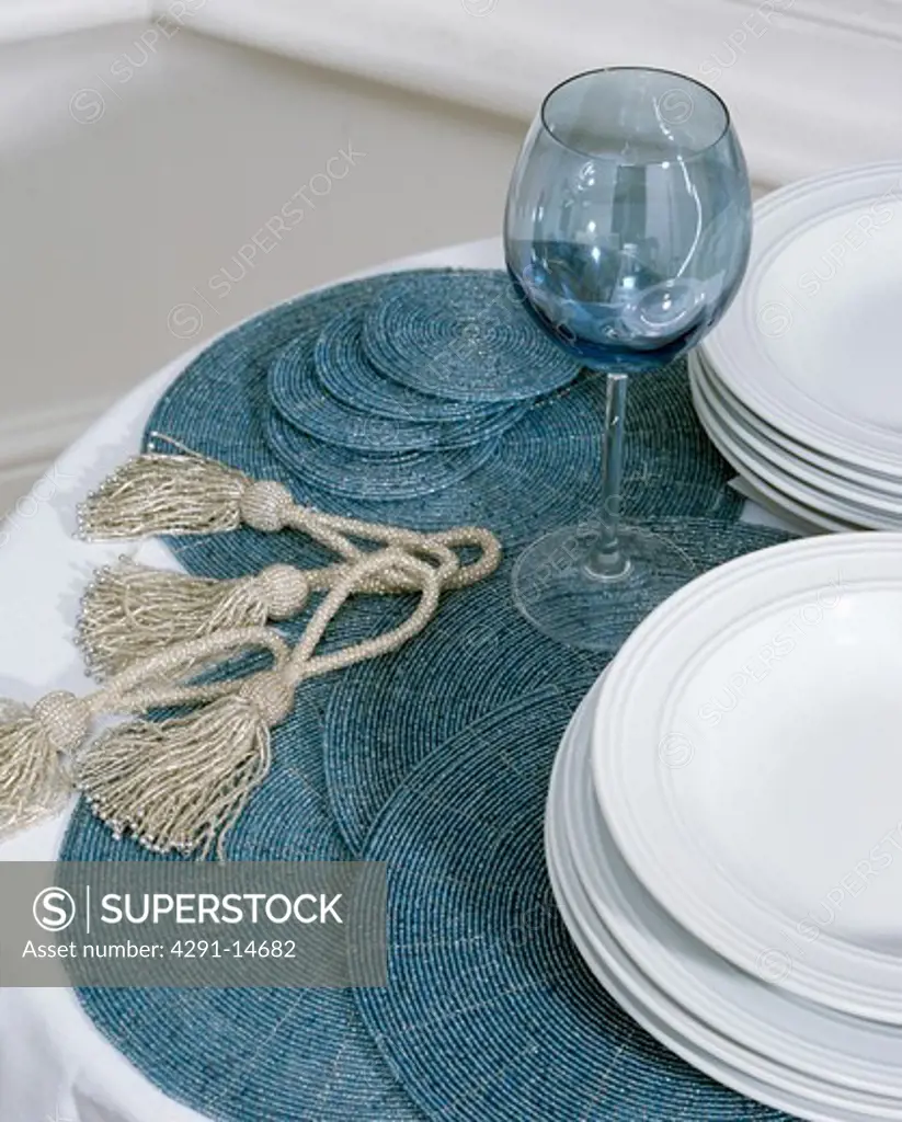 Close-up of circular blue beaded place-mats with beige tassels on table with white plates and blue wine-glass