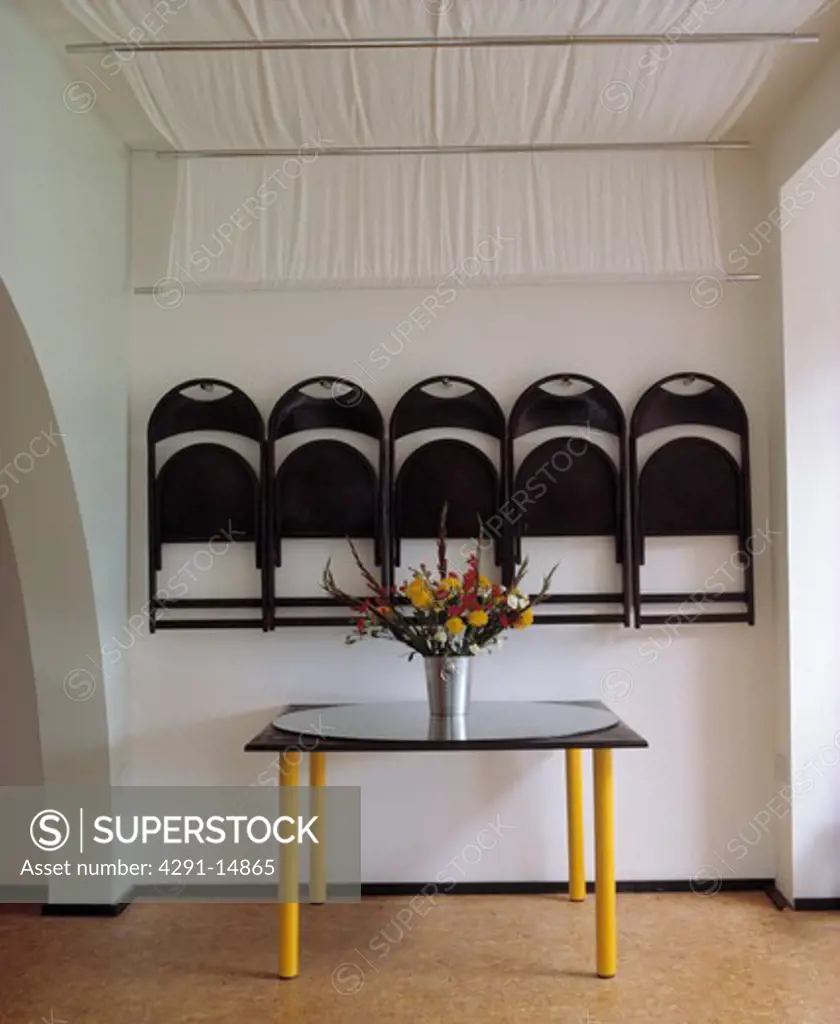 Black folding chairs stored on wall below white ceiling blind in small modern dining room with black and yellow table