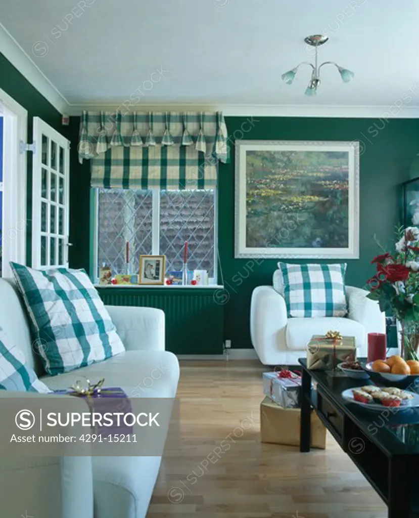 Green checked cushions on white sofa and armchair in dark green living room with green checked blind and wooden floor