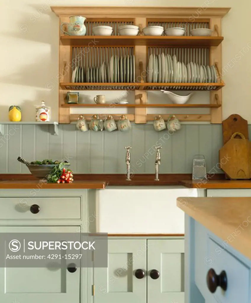 Wooden plate rack above white Belfast sink in kitchen with pale green fitted units