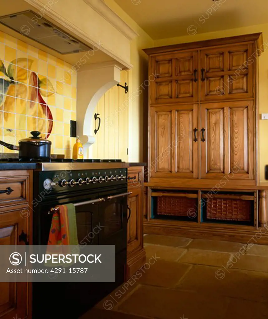 Chestnut-wood cupboard concealing fridge-freezer in yellow kitchen with black range oven and flagstone floor