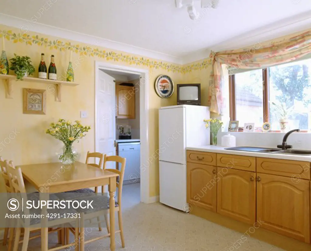 Small television on top of fridge freezer in pastel yellow kitchen dining room with fitted units and wooden table
