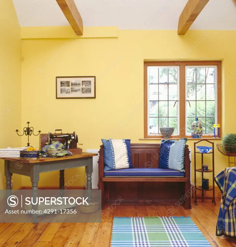 Sewing machine on table beside wooden settle with blue cushions below window in pastel yellow workroom